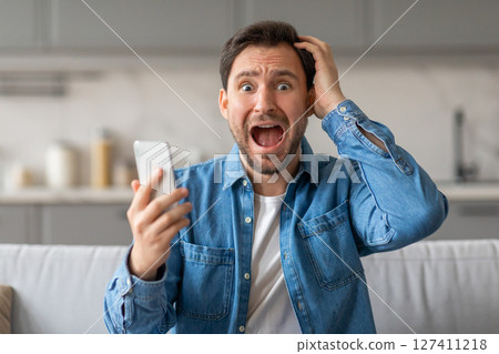 A man in a blue button-down shirt sits on a couch, looking shocked while holding a smartphone in his left hand and holding his right hand to his head. His mouth is open in surprise. A man in a blue button-down shirt sits on a couch, looking shocked while holding a smartphone in his left hand and holding his right hand to his head. His mouth is open in surprise. 127411218