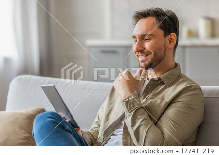 A man is sitting on a white couch in a living room, using a tablet. He is dressed in a tan button down shirt and blue jeans, and has a relaxed expression on his face, copy space A man is sitting on a white couch in a living room, using a tablet. He is dressed in a tan button down shirt and blue jeans, and has a relaxed expression on his face, copy space 127411219