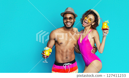 Black man and woman are smiling and enjoying summer cocktails in front of a blue background. The man is wearing sunglasses and a hat while the woman is wearing sunglasses and a swimsuit. 127411233