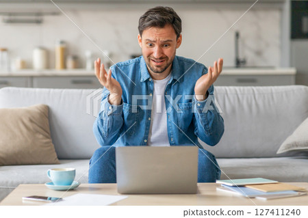 A man sits on a light-colored couch with his hands up in the air looking at a laptop in disbelief. His eyebrows are raised, and his mouth is slightly open in shock. He is wearing a blue denim shirt. 127411240