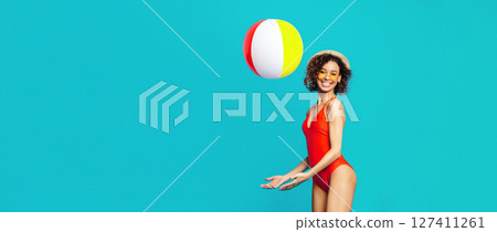 African American young woman in a red swimsuit and a straw hat smiles playfully as she catches a beach ball in a studio with a blue backdrop. The beach ball is red, yellow, and white. African American young woman in a red swimsuit and a straw hat smiles playfully as she catches a beach ball in a studio with a blue backdrop. The beach ball is red, yellow, and white. 127411261