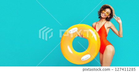 Cute African American lady in red swimsuit, sunglasses and summer hat holding inflatable ring and waving at camera, isolated on blue background, copy space Cute African American lady in red swimsuit, sunglasses and summer hat holding inflatable ring and waving at camera, isolated on blue background, copy space 127411285