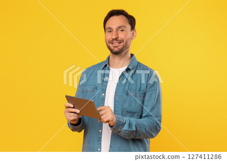 A man in a blue denim shirt stands in front of a yellow background. He is smiling and holding a tablet in his hands. 127411286
