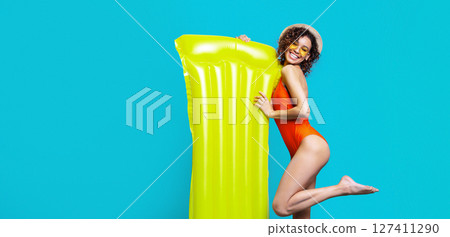 African American young woman in a red swimsuit, sunglasses, and a straw hat smiles while holding a large yellow inflatable raft against a blue background African American young woman in a red swimsuit, sunglasses, and a straw hat smiles while holding a large yellow inflatable raft against a blue background 127411290