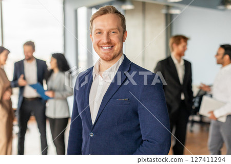 A man in a blue suit and white shirt stands in an office setting, smiling confidently. He is surrounded by colleagues, who are talking and working in the background. The office is bright and modern 127411294