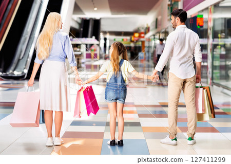 A family of three, a mother, father, and young daughter, walks together through a shopping mall with bags in hand. They are walking away from the camera, holding hands, and looking forward. 127411299