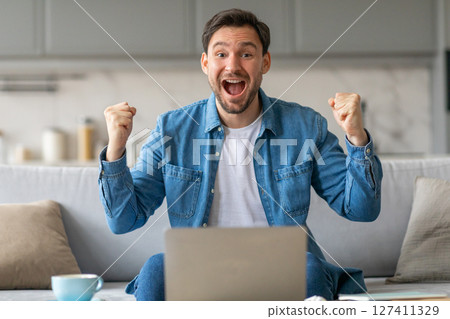 A man with a wide, joyful smile sits on a couch, his hands raised in victory. He is wearing a blue denim shirt and is seated in front of a laptop computer. A man with a wide, joyful smile sits on a couch, his hands raised in victory. He is wearing a blue denim shirt and is seated in front of a laptop computer. 127411329