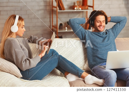 Relaxed Couple In Earphones Using Laptop And Tablet Computers Sitting On Couch Indoor. Weekend At Home. Relaxed Couple In Earphones Using Laptop And Tablet Computers Sitting On Couch Indoor. Weekend At Home. 127411330