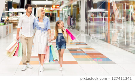 A happy family of three walks through a modern mall, carrying multiple shopping bags. Their daughter is wearing a blue denim overall and a pink shirt. A happy family of three walks through a modern mall, carrying multiple shopping bags. Their daughter is wearing a blue denim overall and a pink shirt. 127411341