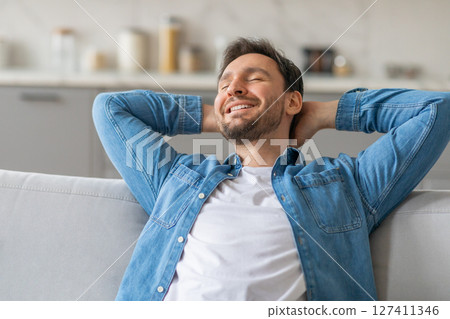 A man wearing a blue denim shirt over a white t-shirt is leaning back on a white couch with his hands clasped behind his head. He has closed eyes and is smiling. 127411346