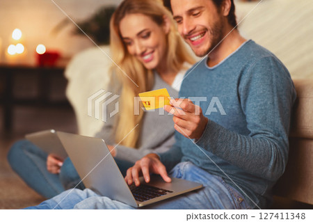 Online Shopping. Happy Couple Ordering On Laptop Paying With Credit Card Sitting On Floor Indoor. Selective Focus Online Shopping. Happy Couple Ordering On Laptop Paying With Credit Card Sitting On Floor Indoor. Selective Focus 127411348