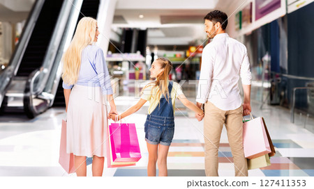 A family of three walks through a mall, holding hands and carrying shopping bags. The parents are smiling as they look towards their daughter, who is walking between them. 127411353