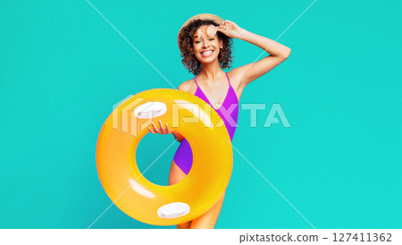 African American woman wearing a purple swimsuit and a straw hat smiles as she holds a large yellow inflatable ring in front of her. The background is a vibrant turquoise color. African American woman wearing a purple swimsuit and a straw hat smiles as she holds a large yellow inflatable ring in front of her. The background is a vibrant turquoise color. 127411362