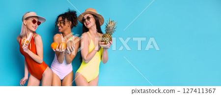 Three women in swimsuits are standing against a bright blue background, holding fruit. One woman holds a pineapple, another holds two oranges, and the third woman holds a banana, copy space 127411367