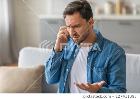 A man with a beard sits on a couch and holds his phone to his ear. He is talking on the phone and seems frustrated, as he has his eyes closed and is gesturing with his free hand A man with a beard sits on a couch and holds his phone to his ear. He is talking on the phone and seems frustrated, as he has his eyes closed and is gesturing with his free hand 127411385