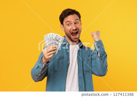 A man in a blue denim shirt and white t-shirt holds a stack of cash in his hand and pumps his fist in the air, celebrating his success. He is looking at the camera with a big smile A man in a blue denim shirt and white t-shirt holds a stack of cash in his hand and pumps his fist in the air, celebrating his success. He is looking at the camera with a big smile 127411386