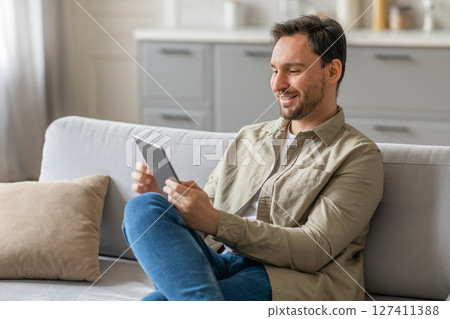 A man in a tan shirt and blue jeans sits on a light gray couch, holding a tablet in both hands. He looks at the tablet with a smile. The couch is in a modern living room with a white, modern kitchen A man in a tan shirt and blue jeans sits on a light gray couch, holding a tablet in both hands. He looks at the tablet with a smile. The couch is in a modern living room with a white, modern kitchen 127411388
