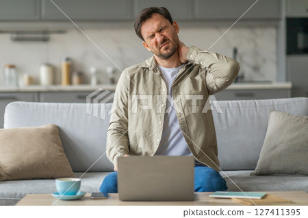 A man sits on a couch, working on a laptop. He is holding his neck and appears to be in pain. The couch is in a living room setting. 127411395