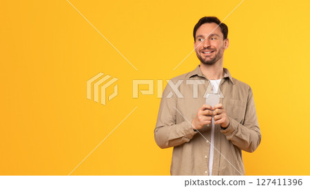 A man in a tan shirt stands against a yellow background, smiling as he looks at copy space while holding a smartphone in his hands. 127411396