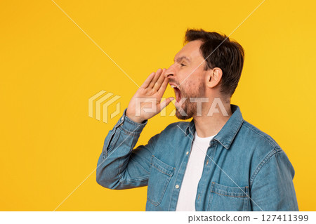 A man with short, dark hair, wearing a light blue button-up shirt, is yelling with his mouth open and a hand cupped near his mouth. He is standing against a plain, bright yellow background. 127411399