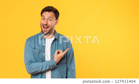 A man in a blue denim shirt smiles and points at copy space while standing in front of a yellow background. 127411404