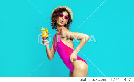 A young African American woman with curly brown hair is wearing a pink swimsuit and sunglasses, holding a cocktail in her hand. She is posing against a solid blue background, smiling at the camera. 127411407