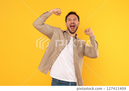 A young man wearing a light brown button-down shirt over a white t-shirt and blue jeans celebrates with raised fists and a wide, excited grin. He is standing against a bright yellow background. 127411409