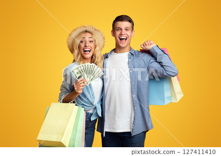 Tax Free Concept. Excited Young Couple Holding Paper Bags And Bunch Of Dollars Happy About Cashback After Successful Shopping Abroad, Panorama With Copy Space 127411410