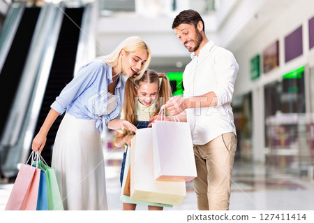 A young girl is standing between her parents, all three looking down at a pile of shopping bags. The girl is smiling, and the parents are smiling as they look at the contents of the bags. 127411414