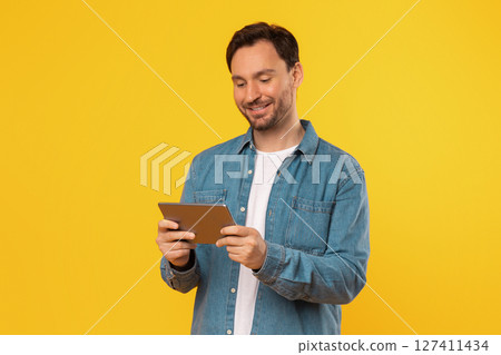 A man wearing a denim shirt stands in front of a bright yellow background, holding a tablet computer in his hands and looking down at the screen. He is smiling as he interacts with the tablet. 127411434