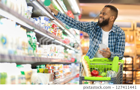 Black Male Buyer Shopping Groceries In Supermarket Taking Dairy Product From Shelf Standing With Shop Cart Indoors. Guy Buys Grocery Choosing Food In Super Market. Empty Space Black Male Buyer Shopping Groceries In Supermarket Taking Dairy Product From Shelf Standing With Shop Cart Indoors. Guy Buys Grocery Choosing Food In Super Market. Empty Space 127411441