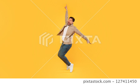 A man with a beard and a tan jacket jumps in the air with his right arm raised, pointing upward. He is wearing blue jeans and white sneakers and is facing the camera. He has a shocked expression A man with a beard and a tan jacket jumps in the air with his right arm raised, pointing upward. He is wearing blue jeans and white sneakers and is facing the camera. He has a shocked expression 127411450