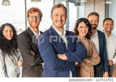 A group of diverse business professionals stands in an office setting, all smiling confidently and looking at the camera. They are wearing professional attire, man in the front has his arms crossed 127411451