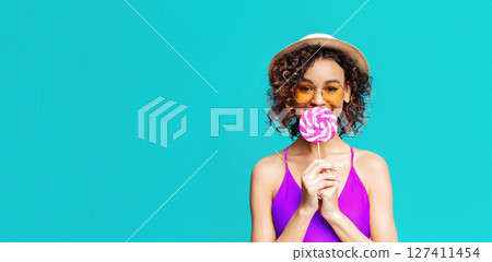 A young woman with curly hair, wearing a purple swimsuit, sunglasses, and a straw hat, smiles while holding a pink and white lollipop in front of her face. She stands against turquoise background. A young woman with curly hair, wearing a purple swimsuit, sunglasses, and a straw hat, smiles while holding a pink and white lollipop in front of her face. She stands against turquoise background. 127411454