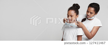 Good african girl helping her little sister to put medical mask on, grey background, copy space 127411467