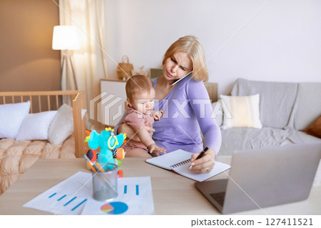 Busy young mother with little baby on her lap having business conversation on mobile phone, taking notes, sitting in front of laptop, home interior, copy space. Working mothers concept 127411521
