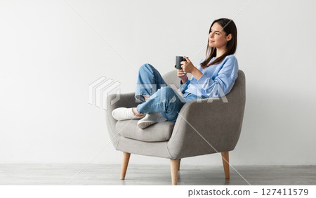 Positive young lady in casual wear sitting in armchair with crossed legs, warming hands on cup of hot coffee, enjoying warm drink against white studio wall, copy space 127411579