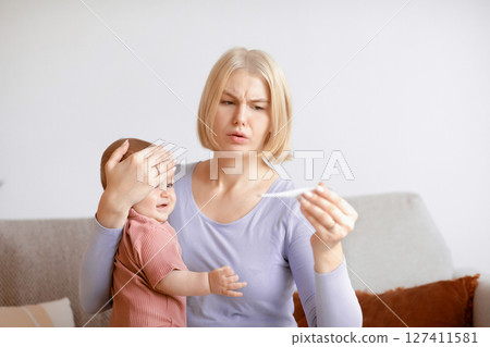 Anxious woman young mother checking temperature to her sick crying baby, using electronic thermometer, home interior. Little kid got fever, troubled mom looking at thermometer. Infant health problems Anxious woman young mother checking temperature to her sick crying baby, using electronic thermometer, home interior. Little kid got fever, troubled mom looking at thermometer. Infant health problems 127411581