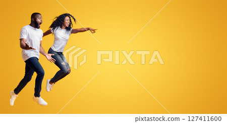 Surprised Black Man Is Looking At Free Space, afro woman is jumping and pointing at empty space, yellow background, panorama 127411600