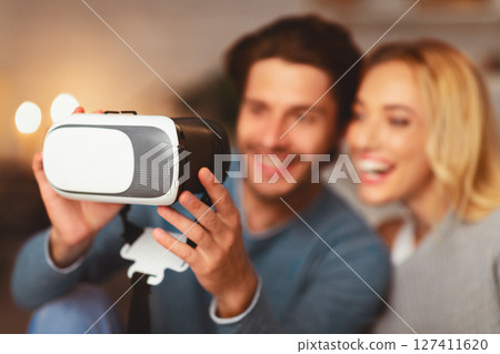 Cheerful Spouses Holding Virtual Reality Headset Relaxing At Home And Playing Videogame. Selective Focus, Shallow Depth 127411620