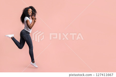 This image shows a young Black woman with curly hair jumping in front of a pink background. She is wearing a white t-shirt and dark blue jeans, and is smiling with her hands raised. This image shows a young Black woman with curly hair jumping in front of a pink background. She is wearing a white t-shirt and dark blue jeans, and is smiling with her hands raised. 127411667