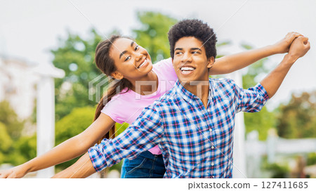 A teen black couple is seen laughing and smiling in an outdoor setting. The girl is holding onto guys hand, pulling him playfully towards her. They appear to be carefree and enjoying each other 127411685