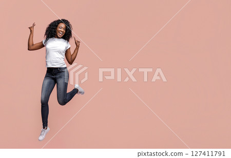 Black woman with dark skin and curly hair jumping in front of a solid pink background. She is wearing a white t-shirt and dark blue jeans. The woman is smiling and looks joyful. 127411791