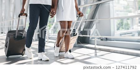 Black couple, a man and a woman, walk side-by-side through an airport terminal while each pulling a suitcase. The photo is taken from a low angle, cropped, copy space 127411792