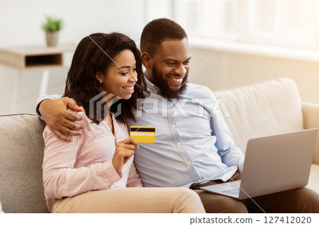 Online Shopping, Ecommerce Concept. Happy young african american couple using laptop and buying clothes or products on internet, paying with debit credit card, sitting on the sofa at home Online Shopping, Ecommerce Concept. Happy young african american couple using laptop and buying clothes or products on internet, paying with debit credit card, sitting on the sofa at home 127412020