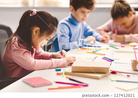 Portrait of small asian girl sitting at table in classroom at primary school or kindergarten, drawing in notebook with colorful pencils. Reopening and return back to school after lockdown 127412021