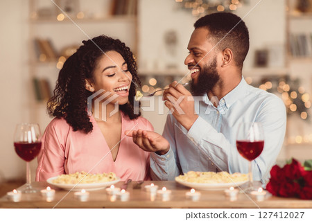 Affectionate young black couple having dinner, guy feeding his girlfriend with pasta. Cute lovers having romantic date, flirting, having fun, celebrating St Valentine's Day or holiday Affectionate young black couple having dinner, guy feeding his girlfriend with pasta. Cute lovers having romantic date, flirting, having fun, celebrating St Valentine's Day or holiday 127412027