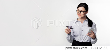 A woman wearing glasses and a white shirt is holding a credit card and a smartphone, smiling as she looks at the screen. A woman wearing glasses and a white shirt is holding a credit card and a smartphone, smiling as she looks at the screen. 127412136