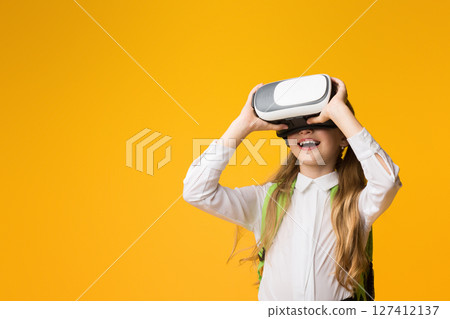 Education Future. Excited Schoolgirl Using VR Headset Experiencing Virtual Reality On Yellow Background. Studio Shot 127412137