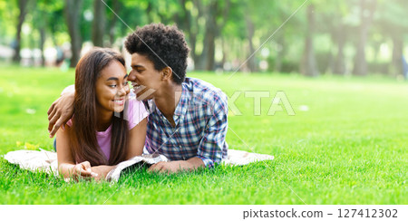 A teen African American couple is lying on a blanket in a park on a sunny day. Guy is lying on his stomach with his arm around his girlfriend, who is looking at him with a smile, copy space A teen African American couple is lying on a blanket in a park on a sunny day. Guy is lying on his stomach with his arm around his girlfriend, who is looking at him with a smile, copy space 127412302